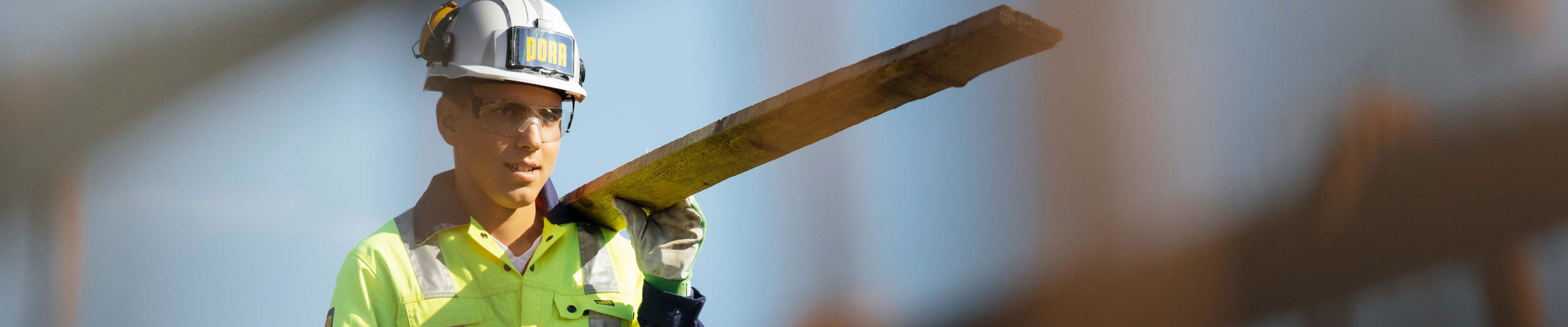 Lehrling Zimmereitechnik mit Schutzausrüstung auf der Baustelle mit Brett in der Hand